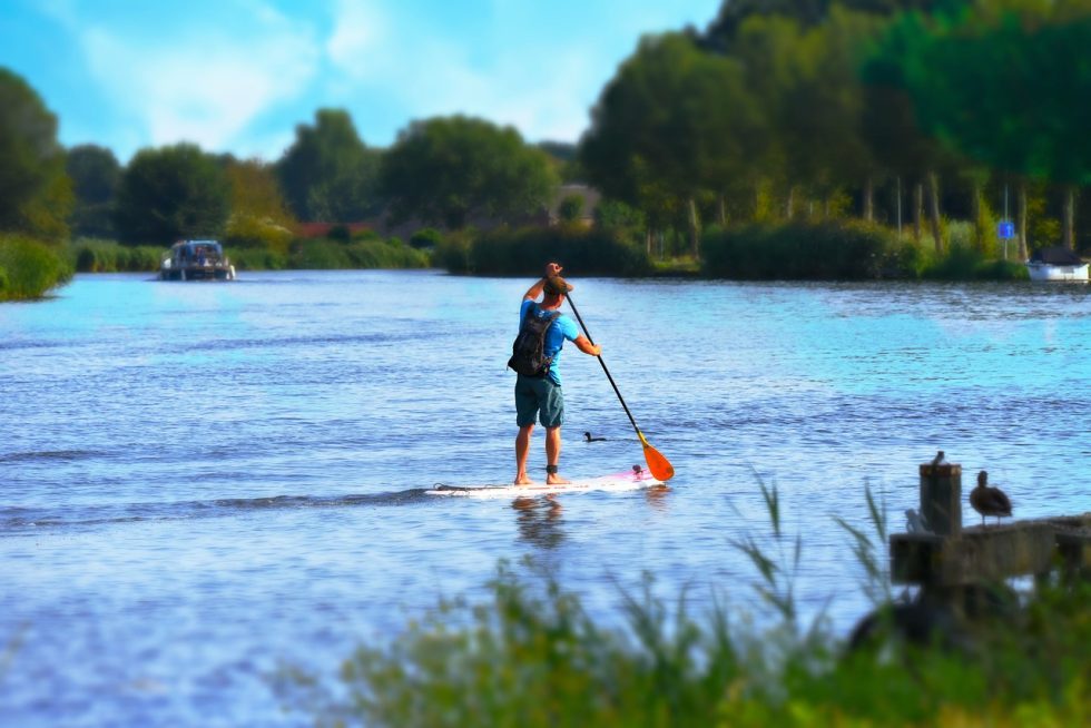 Man met rugzak op aan het SUPPEN op het water met bomen en een boot in de achtergrond